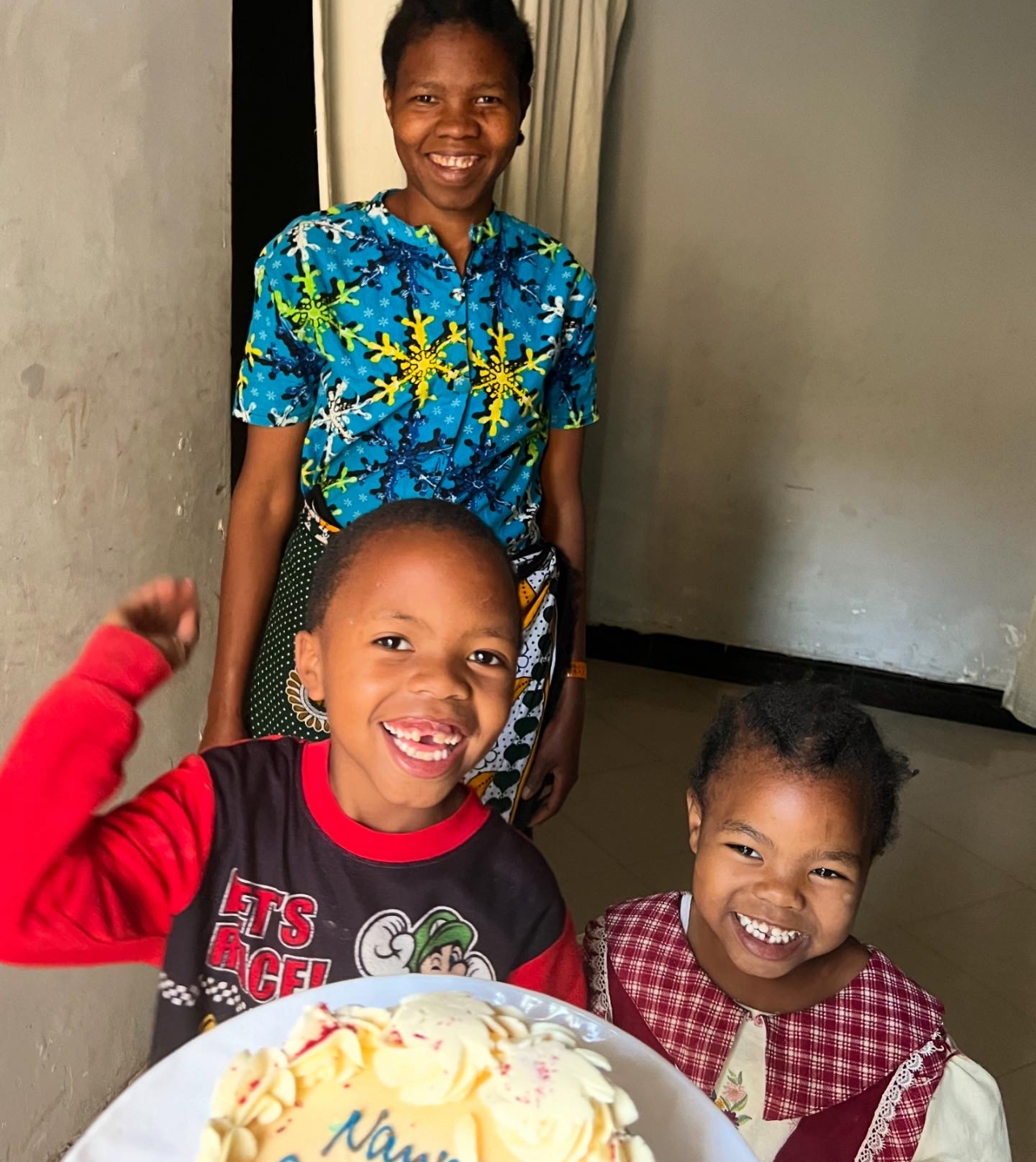Two children smiling beside a decorated cake at home, with an adult standing behind them.